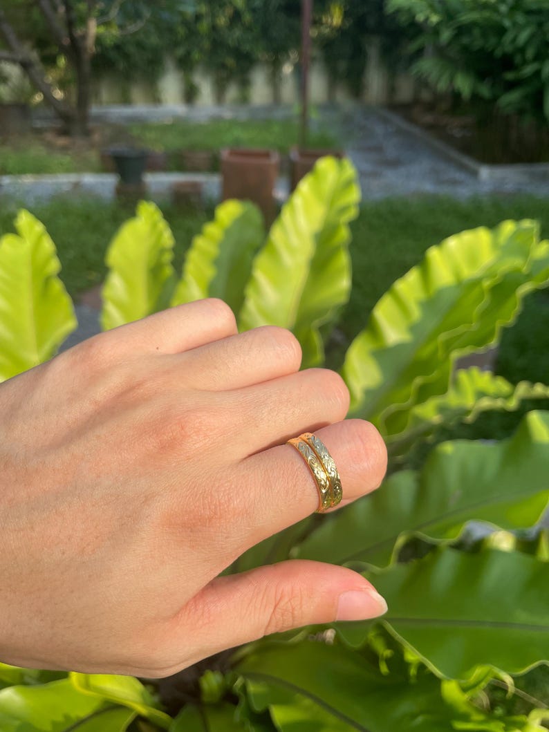 May include: A gold ring with an engraved floral pattern. The ring is worn on a finger, with a blurred background of green foliage. The ring's design features a repeating pattern of leaves and flowers.