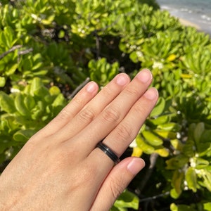 K&ouml;nnte beinhalten: Nahaufnahme einer Hand, die zwei schwarze Ringe tr&auml;gt. Die Ringe sind schlicht und elegant, mit einer matten Oberfl&auml;che. Die Hand ruht auf einem Bett aus gr&uuml;nem Laub.