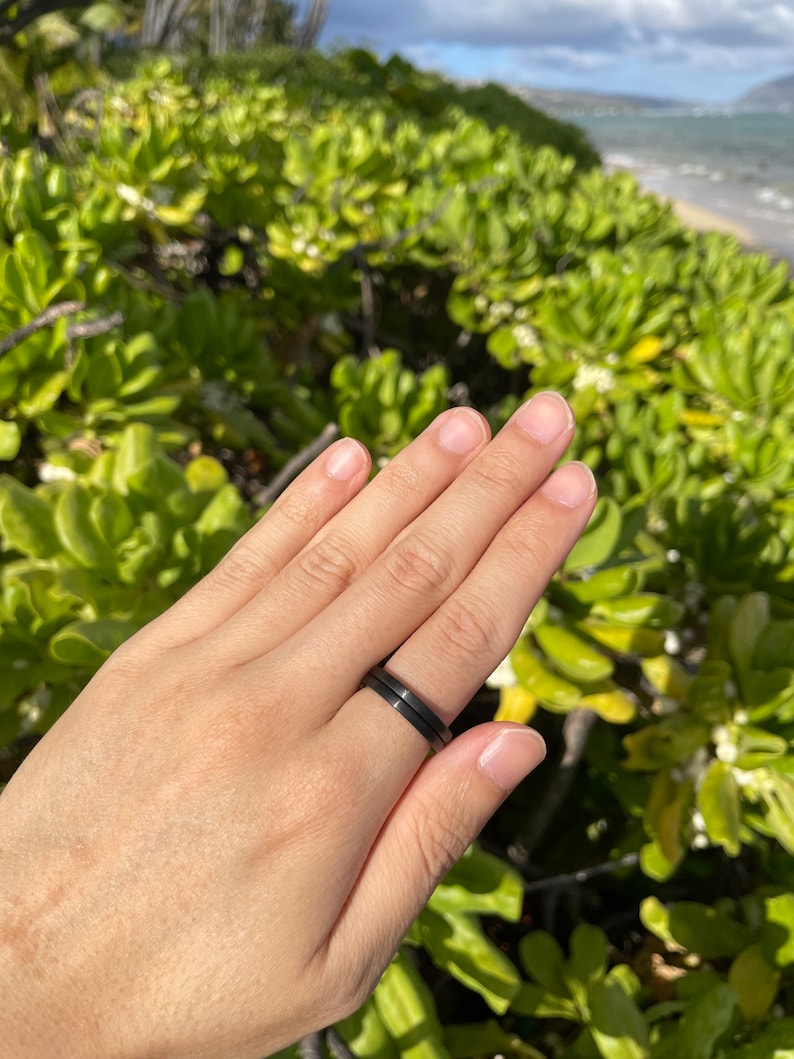 K&ouml;nnte beinhalten: Nahaufnahme einer Hand, die zwei schwarze Ringe tr&auml;gt. Die Ringe sind schlicht und elegant, mit einer matten Oberfl&auml;che. Die Hand ruht auf einem Bett aus gr&uuml;nem Laub.