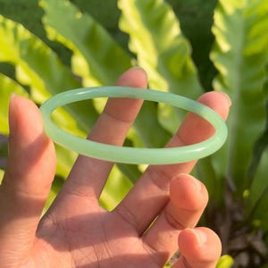 May include: A pale green jade bangle bracelet. The circular bracelet is held in a hand, with green foliage in the background. The jade has a translucent quality, and the overall aesthetic is natural and elegant.