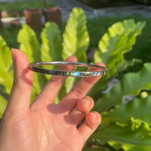 Puede incluir: Un brazalete plateado con una fina incrustación iridiscente. El brazalete está siendo sostenido por una mano.