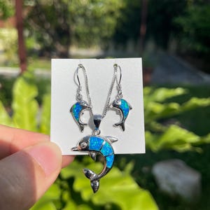 May include: A silver dolphin pendant necklace and matching earrings set. The dolphins have blue opal-like inlays. The jewellery is displayed on a white card against a blurred green background.