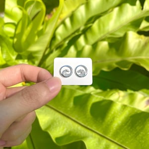 May include: A pair of silver wave design stud earrings on a white card. The earrings are circular with a swirling wave pattern. The background features lush green foliage.