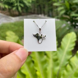 Puede incluir: Un collar con colgante plateado con diseño de manta raya. El colgante cuelga de una fina cadena y se presenta sobre una tarjeta blanca. El fondo está borroso, mostrando follaje verde.