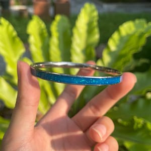 Puede incluir: Una pulsera de brazalete plateada con una vibrante incrustación turquesa. La pulsera tiene un acabado liso y pulido y una banda de material azul-verdoso brillante. La joya se sostiene sobre un fondo de follaje verde.