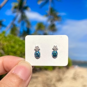Op de afbeelding: Een paar zilverkleurige oorstekers in de vorm van ananassen. De ananaslichamen zijn een mozaïek van blauw en blauwgroen, met een getextureerde zilveren kroon. De oorbellen worden op een witte kaart getoond.