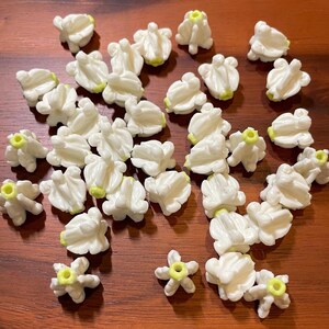 May include: A collection of small, white, flower-shaped beads with yellow centers. The beads are scattered on a wooden surface, showcasing their delicate design and potential for crafting projects. The beads are approximately 0.5 inches in diameter.