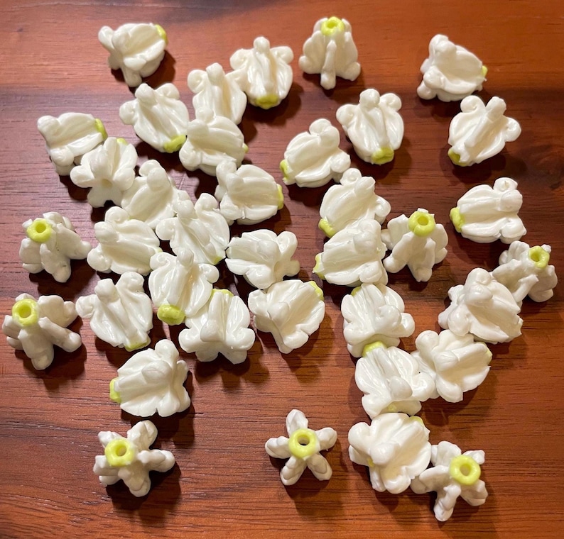 May include: A collection of small, white, flower-shaped beads with yellow centers. The beads are scattered on a wooden surface, showcasing their delicate design and potential for crafting projects. The beads are approximately 0.5 inches in diameter.