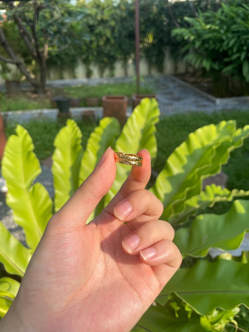 May include: A gold-colored ring with a textured design. The ring is held up in front of a background of green foliage. The ring appears to be made of metal and has a detailed pattern.