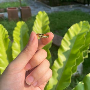 May include: A gold ring with an engraved floral pattern. The ring is held in front of a background of green foliage. The ring is a small piece of jewelry, likely worn on a finger.