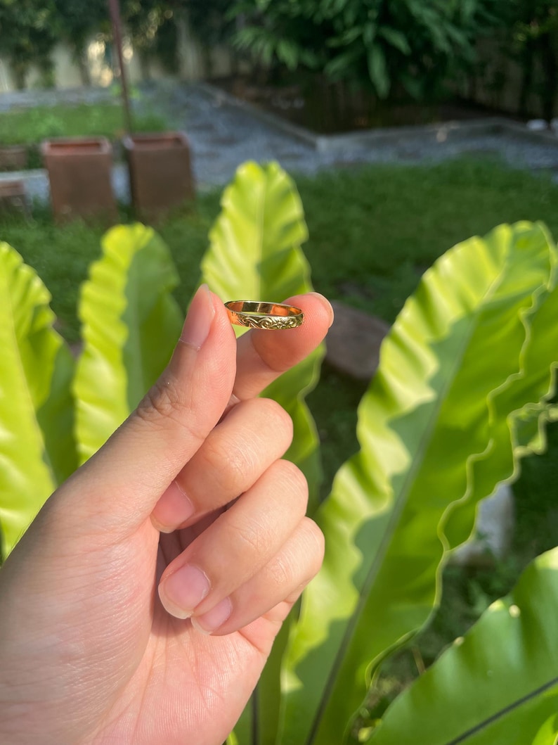 May include: A gold ring with an engraved floral pattern. The ring is held in front of a background of green foliage. The ring is a small piece of jewelry, likely worn on a finger.