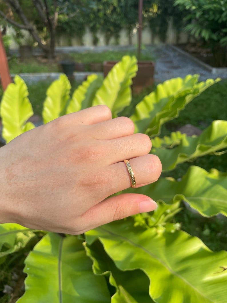 May include: A gold ring with a textured surface is worn on a finger. The ring appears to be a band style, with a repeating pattern. The background features green foliage and natural light.