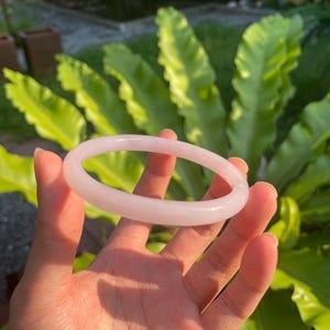 May include: A light pink jade bangle bracelet. The smooth, polished bracelet is circular and held in a hand, with green foliage in the background. The bangle is a soft, translucent color.