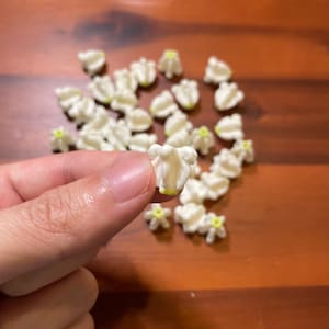 May include: A collection of small, white, bell-shaped flower beads with subtle yellow centers. Each bead features a textured, multi-petaled design resembling a lily of the valley. The beads are scattered, with one held up to show detail.