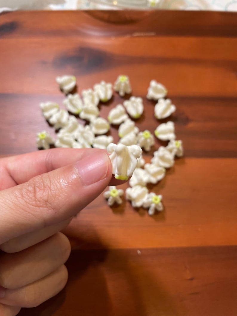 May include: A collection of small, white, bell-shaped flower beads with subtle yellow centers. Each bead features a textured, multi-petaled design resembling a lily of the valley. The beads are scattered, with one held up to show detail.