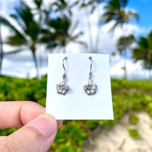 May include: Silver-colored hibiscus flower earrings. The earrings are displayed on a white card, held against a backdrop of palm trees and a blue sky. The earrings have a hook closure.