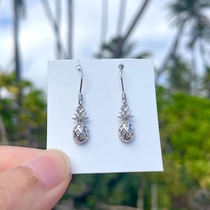 Pode incluir: Um par de brincos de abacaxi prateados. Cada brinco apresenta um pingente de abacaxi detalhado suspenso de um gancho. Os brincos são exibidos em um cartão branco, com um fundo desfocado verde e azul.