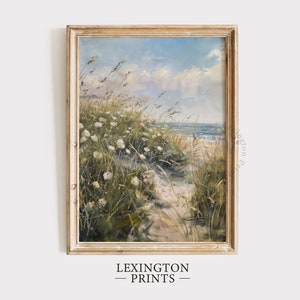 Puede incluir: Una pintura enmarcada que representa una escena de playa con flores blancas y hierbas altas en primer plano, un camino de arena que conduce al océano y un cielo azul con nubes blancas.