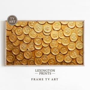 May include: A framed print of a gold painting of four-leaf clovers. The painting is a close-up of many clovers, all overlapping each other. The clovers are a shiny gold color and have a textured appearance. The frame is a simple, light wood color.