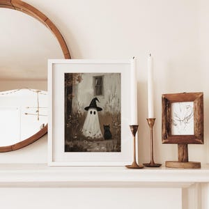 Puede incluir: Una impresi&oacute;n art&iacute;stica enmarcada con tem&aacute;tica de Halloween que presenta un fantasma blanco con un sombrero de bruja negro, con un gato negro a su lado. La obra de arte est&aacute; ambientada sobre un fondo sepia con una casa y follaje. La impresi&oacute;n est&aacute; en una repisa blanca.