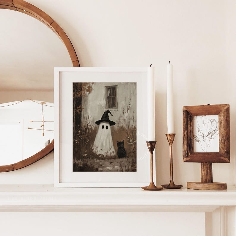 Puede incluir: Una impresi&oacute;n art&iacute;stica enmarcada con tem&aacute;tica de Halloween que presenta un fantasma blanco con un sombrero de bruja negro, con un gato negro a su lado. La obra de arte est&aacute; ambientada sobre un fondo sepia con una casa y follaje. La impresi&oacute;n est&aacute; en una repisa blanca.