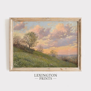 May include: A framed painting of a landscape with a rolling green hill, a few trees, and a cloudy sky with a pink and orange sunset. The painting is in a light wood frame.