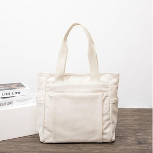 May include: A cream-colored canvas tote bag featuring two sturdy top handles, a large rectangular front pocket, and two side pockets. The bag has a structured rectangular shape with visible stitching along the seams and a plain fabric exterior.