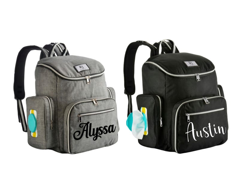 May include: Two diaper backpacks, one grey and one black, with multiple pockets and a top handle. The grey backpack has the name "Alyssa" in cursive, and the black backpack has the name "Austin". Both have a side pocket for wipes.
