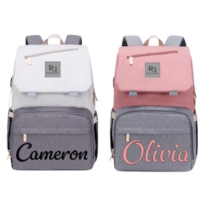 May include: Two diaper backpacks, one white and gray, the other pink and gray. Each has a flap closure, a front pocket, and a gold zipper. The white backpack has the name "Cameron" in black script, and the pink backpack has the name "Olivia" in pink script.