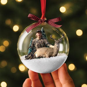 May include: A clear glass Christmas bauble with a red and black tartan ribbon. Inside the bauble is a person in camouflage clothing with a deer and small Christmas trees on a bed of white snow.