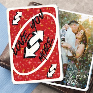 Peut inclure: Deux cartes photo personnalisées. L'une est une carte Uno rouge avec les mots "Love You More" et des flèches inversées. L'autre carte présente une photo d'un couple avec le texte "& Eve 2025".