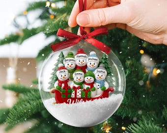 May include: A clear Christmas ornament with a family of snowmen inside. The snowmen are wearing hats and scarves, and are in a red sleigh with the word "Wish". The ornament is held by a red ribbon.