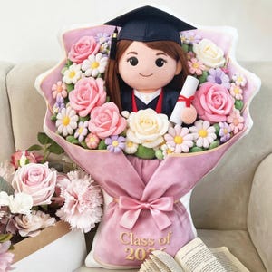 Op de afbeelding: Een zachte afstudeerpop met een hoed en toga, omringd door een boeket roze, witte en gele bloemen. Het boeket is verpakt in roze stof en vastgebonden met een strik. De tekst "Class of 2023" is zichtbaar.
