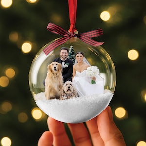 May include: A clear glass Christmas bauble containing a wedding photograph. The image shows a bride, groom, and two dogs, set in a snowy scene. The bauble is suspended by a red and black tartan ribbon.