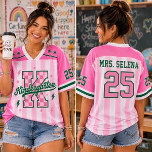 May include: A pink and white striped V-neck jersey featuring a large checkered letter K with the word Kindergarten in green script. The back displays the text MRS. SELENA and the number 25 in green. The sleeves have green and white stripes.