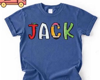 May include: A blue short-sleeve t-shirt featuring the name JACK printed in large, colorful letters. The J is a red apple, the A is white lined paper, the C is a yellow pencil, and the K is a green ruler. The top left corner shows a small truck icon with the text NEXT DAY.