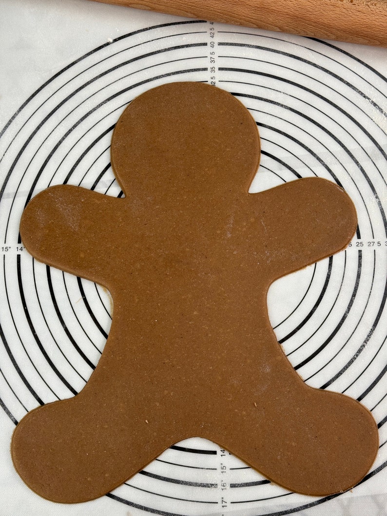 May include: A brown gingerbread cookie in the shape of a gingerbread man on a white baking mat with black concentric circles.