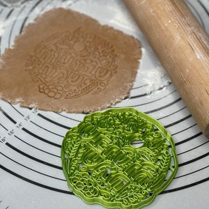 May include: A green cookie cutter featuring an intricate design of a floral wreath and the word "Happy" in a decorative font. The cutter is on a white surface with a rolling pin and a piece of dough.