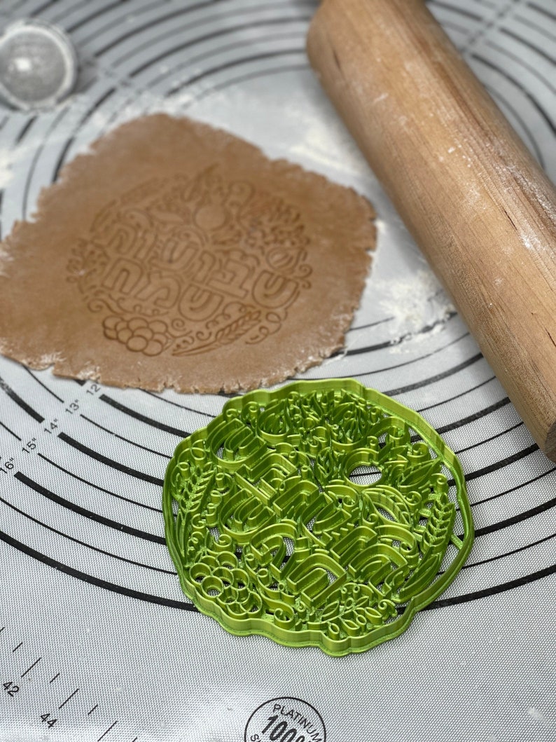 May include: A green cookie cutter featuring an intricate design of a floral wreath and the word "Happy" in a decorative font. The cutter is on a white surface with a rolling pin and a piece of dough.