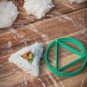 May include: A green plastic triangle-shaped sushi mold on a wooden surface with a clear plastic wrap. There are four balls of white rice and one triangle-shaped sushi roll with fillings.