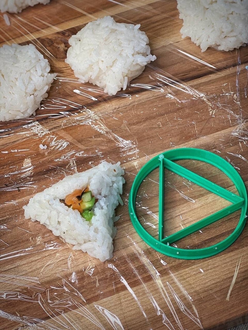 May include: A green plastic triangle-shaped sushi mold on a wooden surface with a clear plastic wrap. There are four balls of white rice and one triangle-shaped sushi roll with fillings.