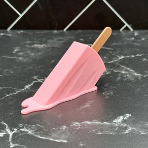 Puede incluir: Un helado de paleta rosa con un palo de madera, derriti&eacute;ndose sobre una superficie de m&aacute;rmol blanco y negro.