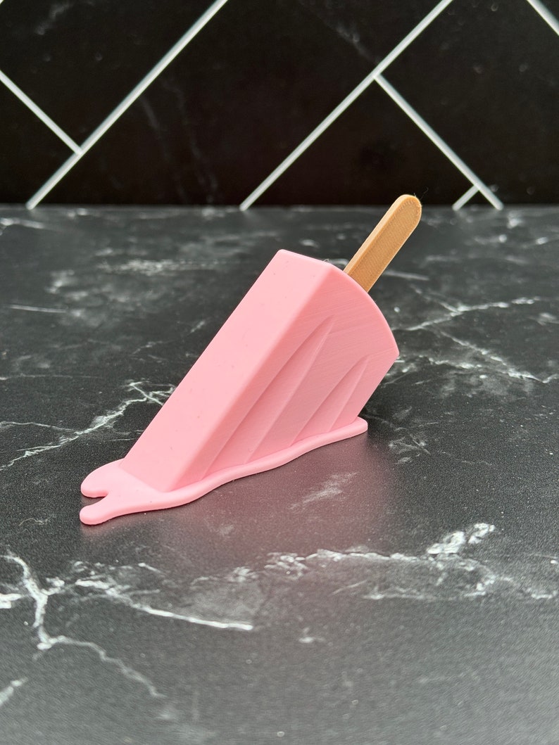 Puede incluir: Un helado de paleta rosa con un palo de madera, derriti&eacute;ndose sobre una superficie de m&aacute;rmol blanco y negro.