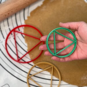 May include: Three round cookie cutters, one red, one green, and one gold, each with a triangle design inside. The cutters are on a white surface with a brown rolling pin and a sheet of dough.