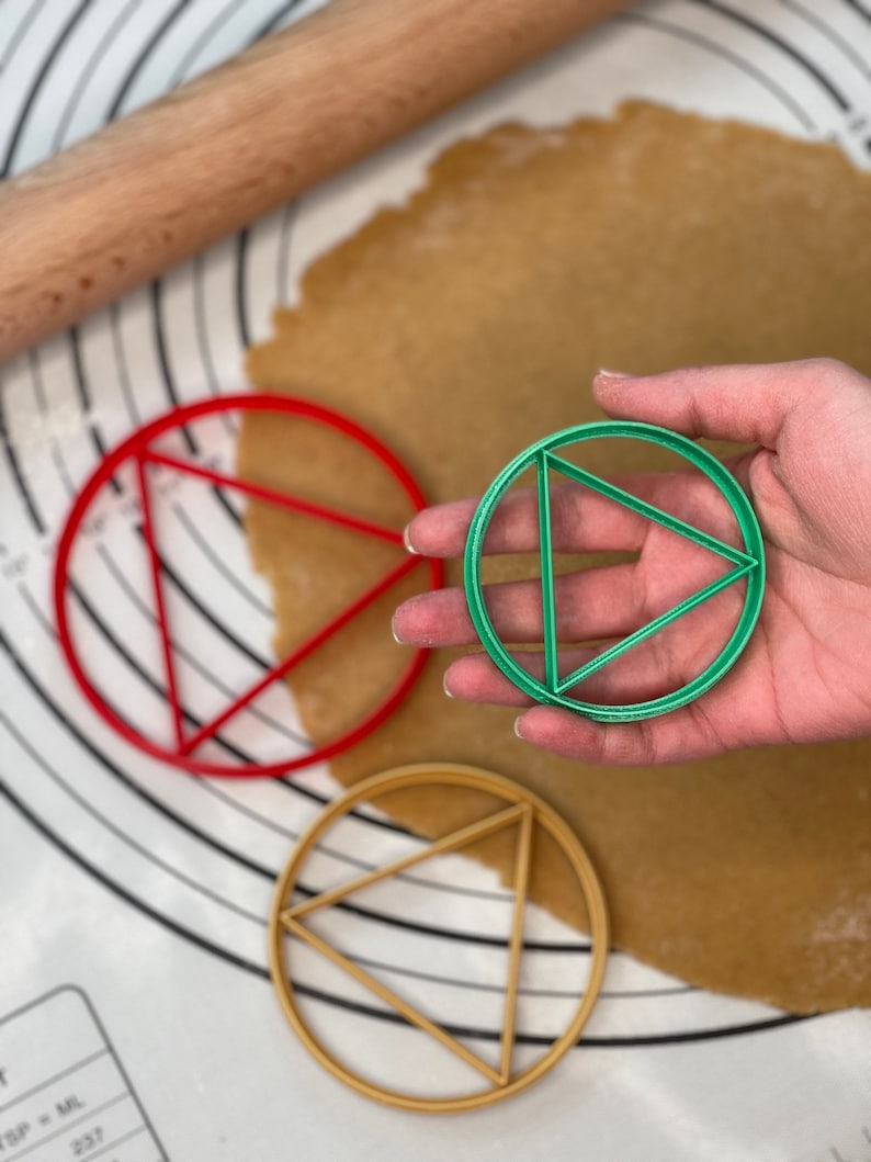 May include: Three round cookie cutters, one red, one green, and one gold, each with a triangle design inside. The cutters are on a white surface with a brown rolling pin and a sheet of dough.