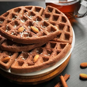 Peut inclure: Une pile de gaufres au chocolat garnies de noix de coco râpée et d'amandes, servies sur une assiette blanche avec une base en bois. Des bâtonnets de cannelle et de l'anis étoilé sont disposés sur une surface noire.