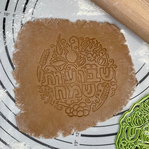 May include: A brown gingerbread cookie dough with a decorative imprint of a pomegranate, grapes, wheat, and the Hebrew words "Shanah Tovah" in a circle. A green plastic cookie cutter with the same design is in the background.