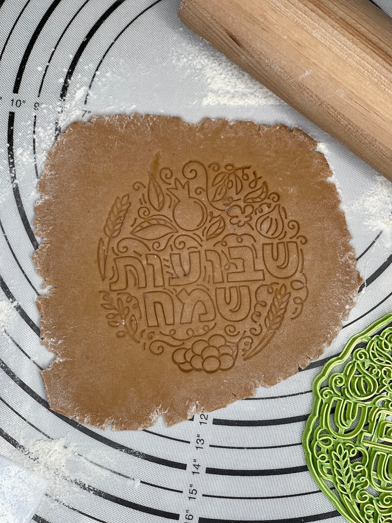 May include: A brown gingerbread cookie dough with a decorative imprint of a pomegranate, grapes, wheat, and the Hebrew words "Shanah Tovah" in a circle. A green plastic cookie cutter with the same design is in the background.