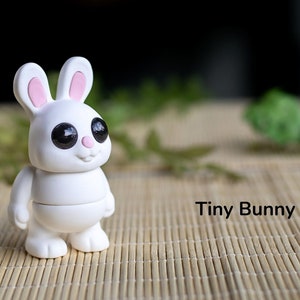 May include: A white ceramic bunny figurine with pink ears and black eyes. The bunny is standing on a woven bamboo mat. The text "Tiny Bunny" is written below the bunny.