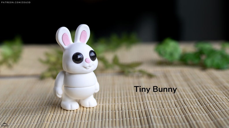 May include: A white ceramic bunny figurine with pink ears and black eyes. The bunny is standing on a woven bamboo mat. The text "Tiny Bunny" is written below the bunny.